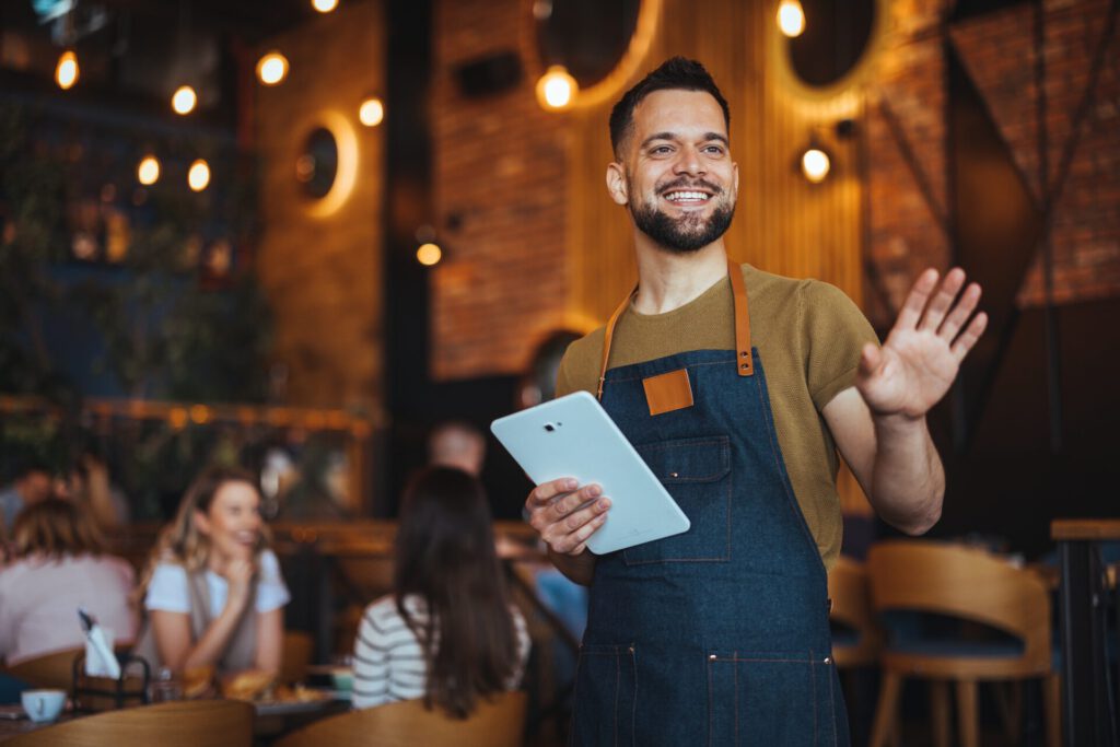 A,Cheerful,Waiter,Welcomes,Guests,While,Holding,A,Tablet,In